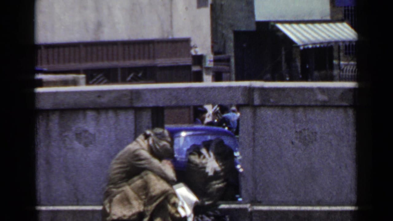 Huddled person wrapped in a blanket beside a blue street object near a low concrete wall.