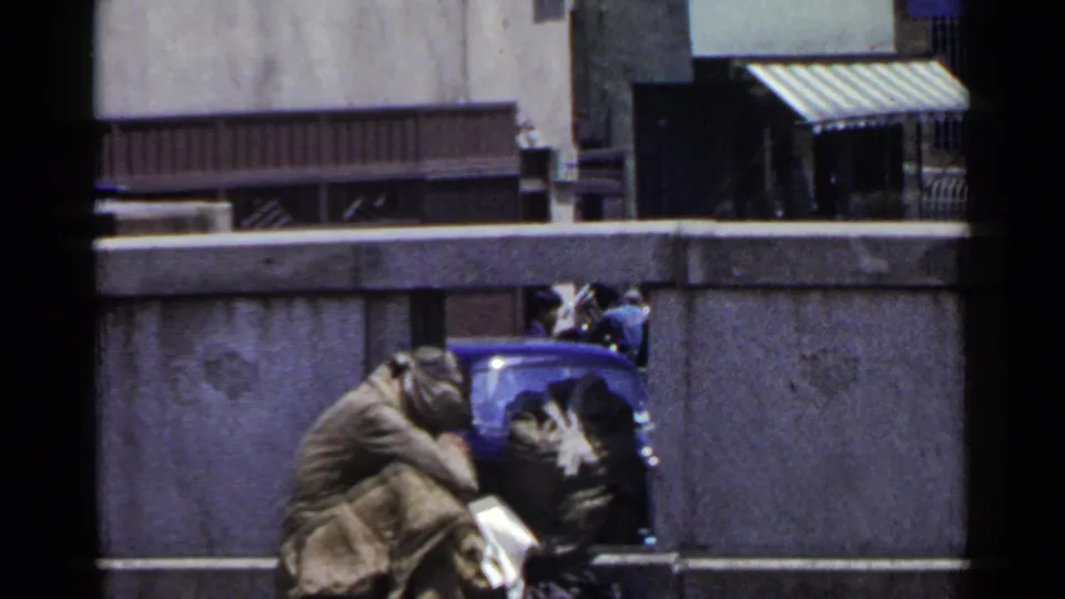 1953 Japan home-movie frame showing a huddled person in a blanket beside a blue object on a city street.