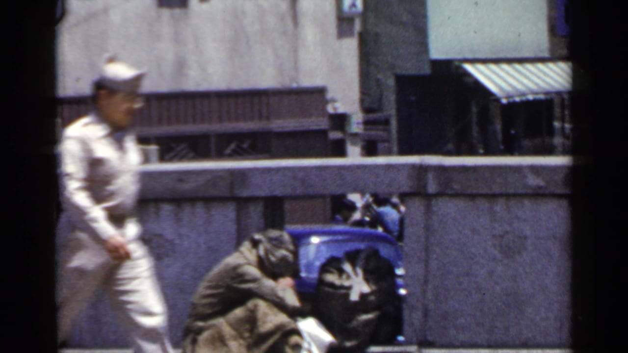 1953 Japan street frame with a light-uniformed passerby crossing in front of a huddled person by a wall.