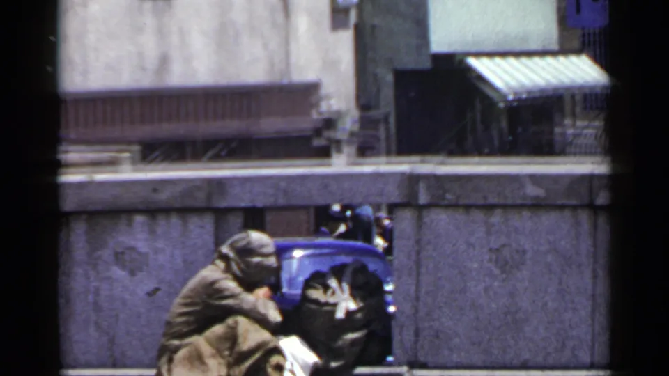 1953 Japan home-movie view of a blanket-wrapped person crouched near a wall on an urban street.