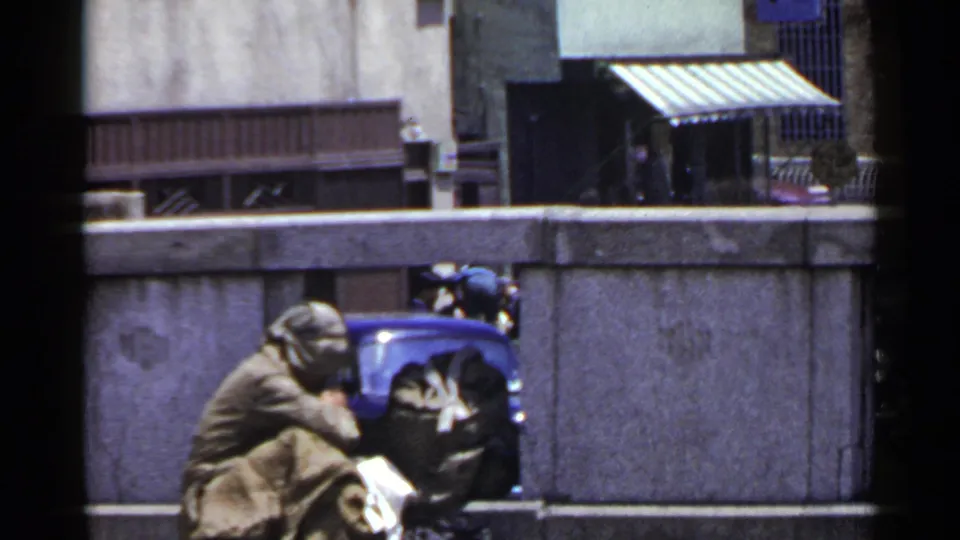 1953 Japan street scene showing a huddled person in a blanket beside a blue object near a concrete wall.