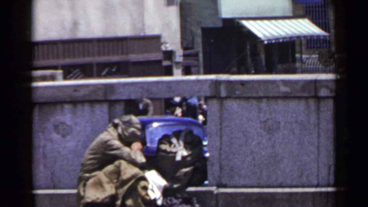In 1953 Japan home-movie footage, a man appears huddled by a concrete wall with bundled belongings beside him.