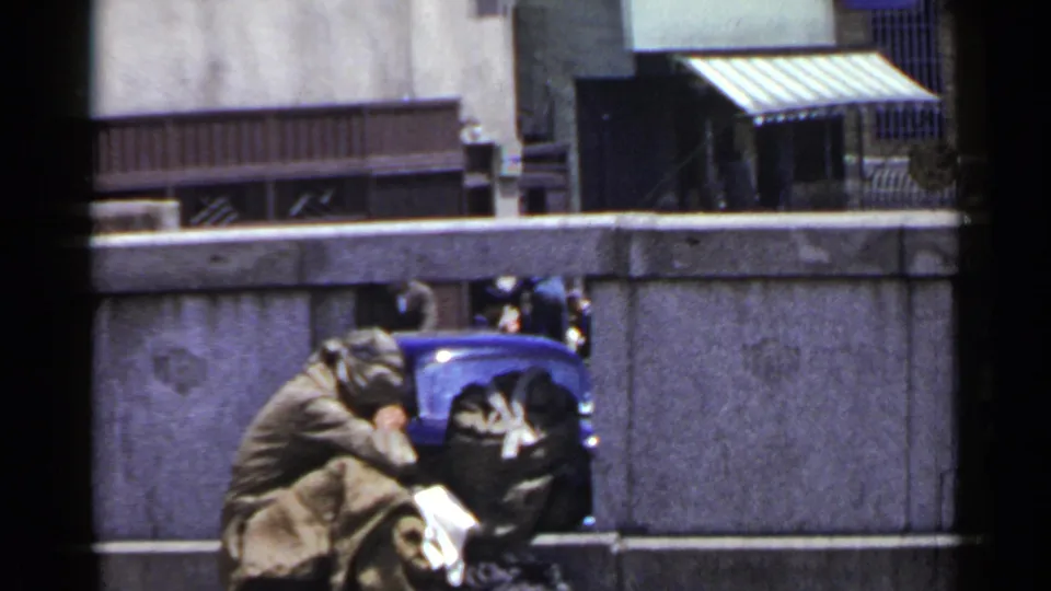 In 1953 Japan home-movie footage, a man appears huddled by a concrete wall with bundled belongings beside him.