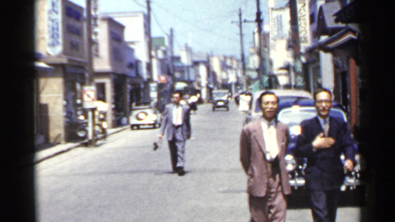 Two men in suits walk along a Japanese urban street in 1953, with vintage cars, storefronts, and vertical signs visible.