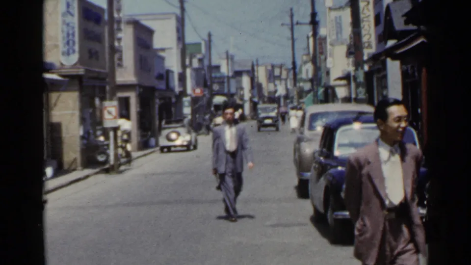 A suited man passes near the camera while another pedestrian walks through a mid-century Japanese street with parked cars and shops.