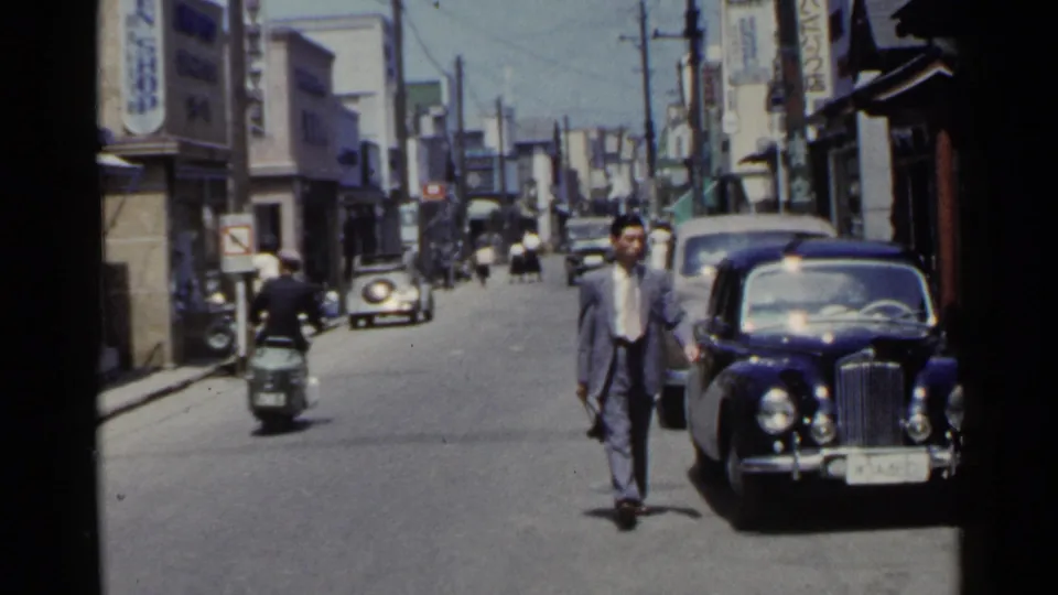 A pedestrian walks near a dark vintage car on a Japanese street in 1953, with a motorcyclist and storefront signs in the background.