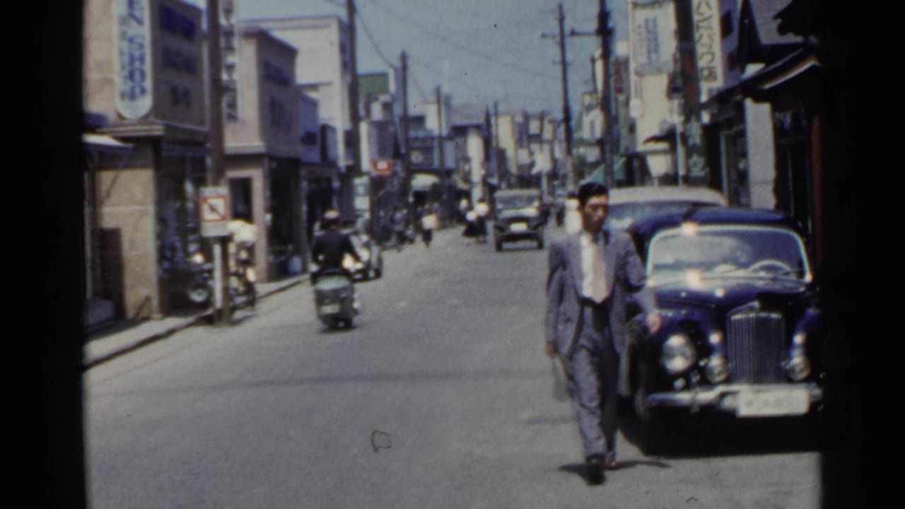 1953 Japan street view with a suited man walking near a dark classic car as scooters and pedestrians move in the background.