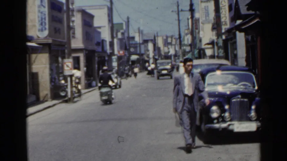 1953 Japan street view with a suited man walking near a dark classic car as scooters and pedestrians move in the background.