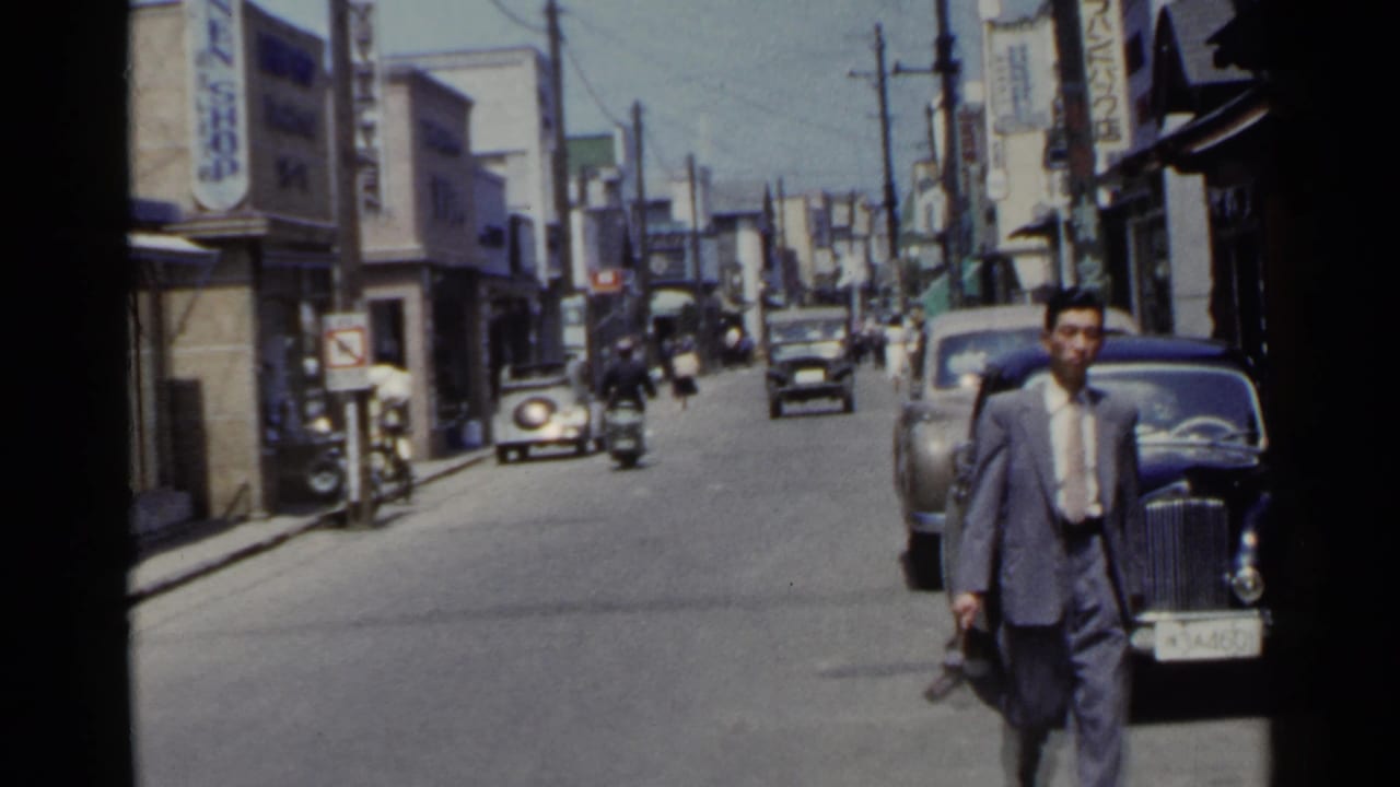 1953 Japan town street with a suited pedestrian close to camera, a dark classic car, and small vehicles moving along the road.