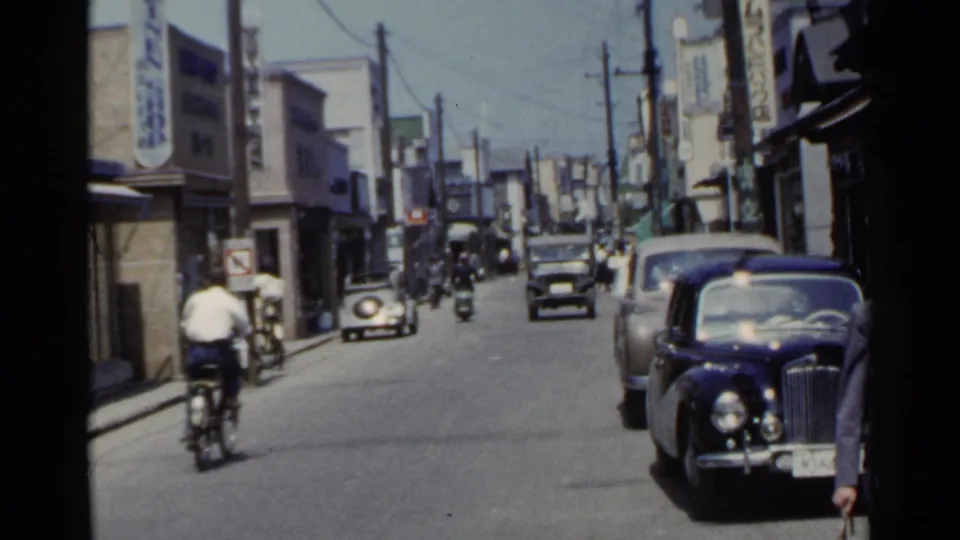 1953 Japan street scene with a cyclist in the foreground, classic cars along the curb, and storefront signs lining the road.