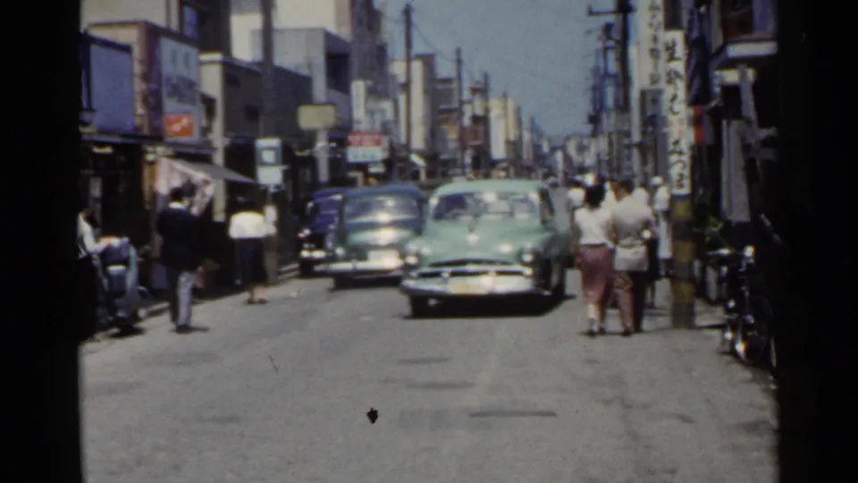1953 Japan street with a pale green mid-century car approaching, pedestrians at right, and shopfront signage along the road.