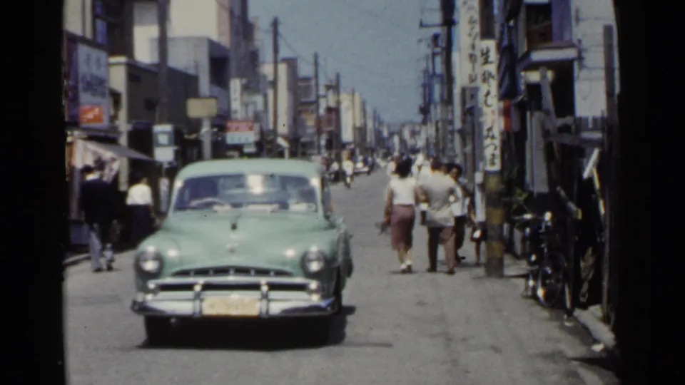 1953 Japan street view with a pale green car near the camera, pedestrians walking away, and vertical shop signs in the background.