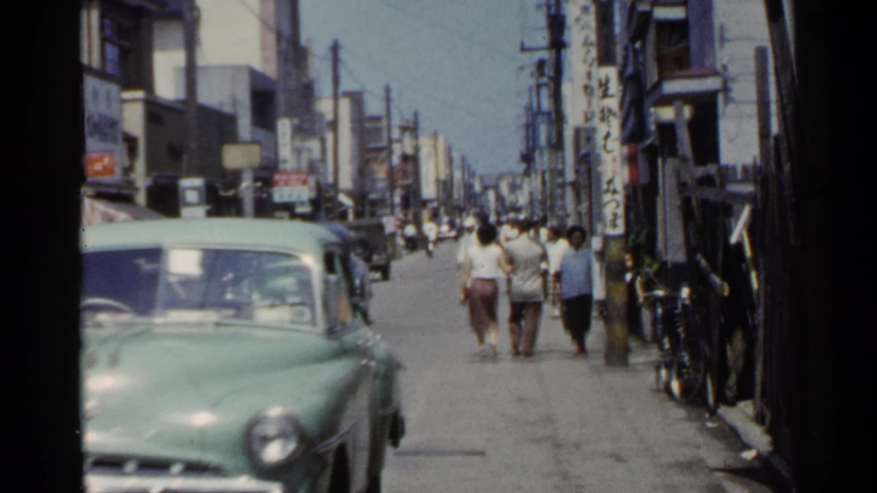 1953 Japan street view with a green sedan, pedestrians, storefronts, overhead wires, and vertical shop signs.
