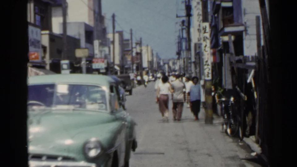1953 Japan street view with a green sedan, pedestrians, storefronts, overhead wires, and vertical shop signs.