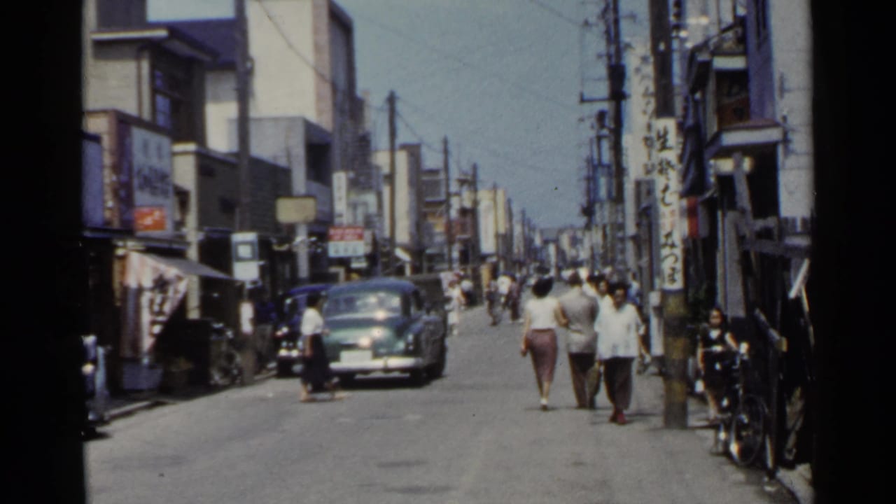 1953 Japan commercial street with pedestrians, a dark car, shop signs, bicycles, and dense overhead wiring.