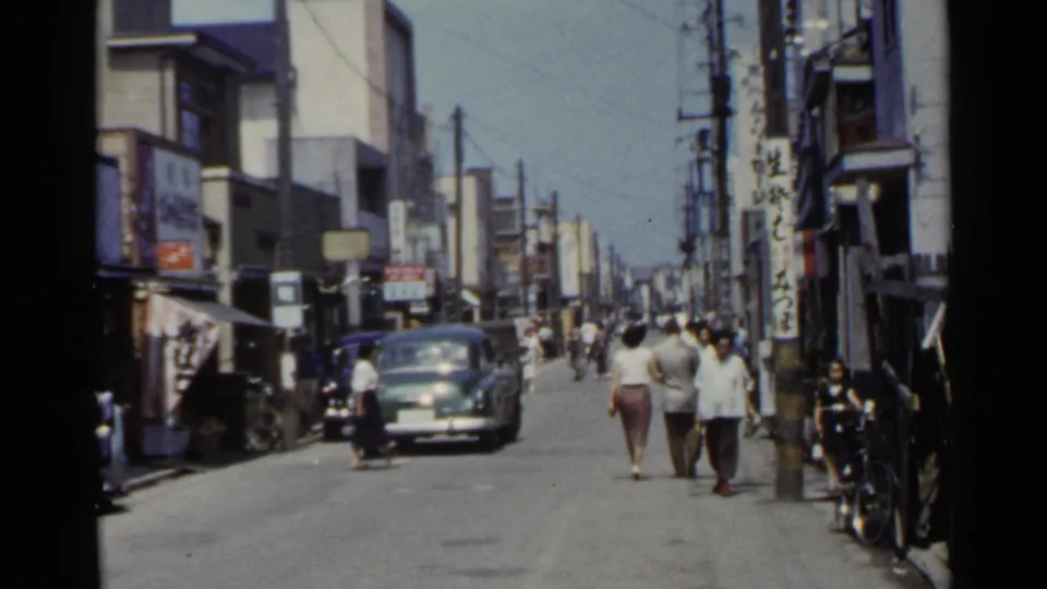 1953 Japan commercial street with pedestrians, a dark car, shop signs, bicycles, and dense overhead wiring.