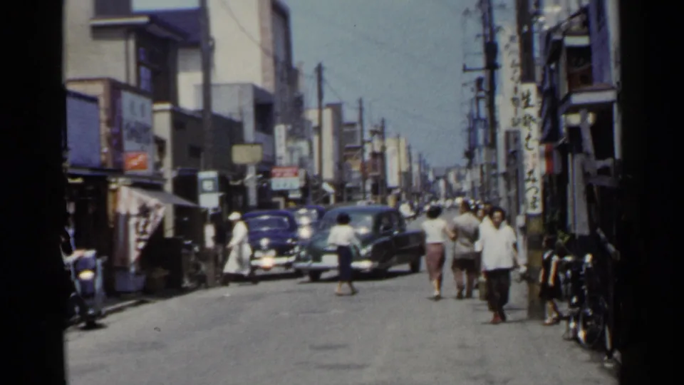 1953 Japan street scene with two vintage cars, walkers, storefront awnings, bicycles, and Japanese signs.