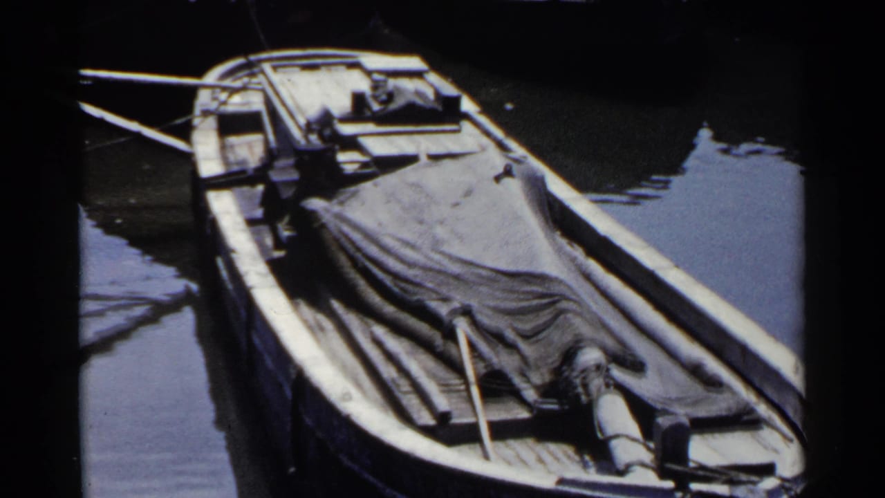 1953 Japan home-movie frame of a tarped wooden boat resting near a dock in still water, with deck details visible.