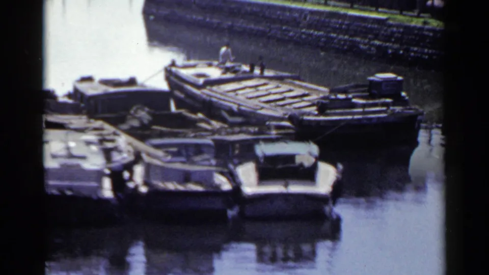 A Japanese harbor canal holds wooden barges and small boats, with one distant person visible on a deck.