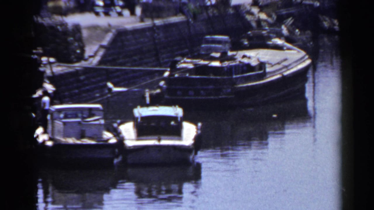 Small boats moored along a canal with a bridge and dock activity in the background.