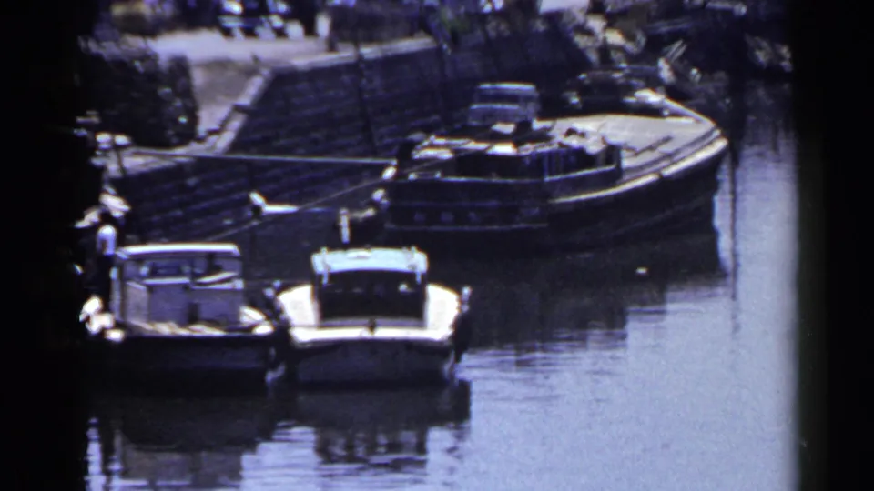 1953 Japan 8mm frame showing small boats tied along a calm waterway, with a bridge, quay, and distant pedestrians behind them.