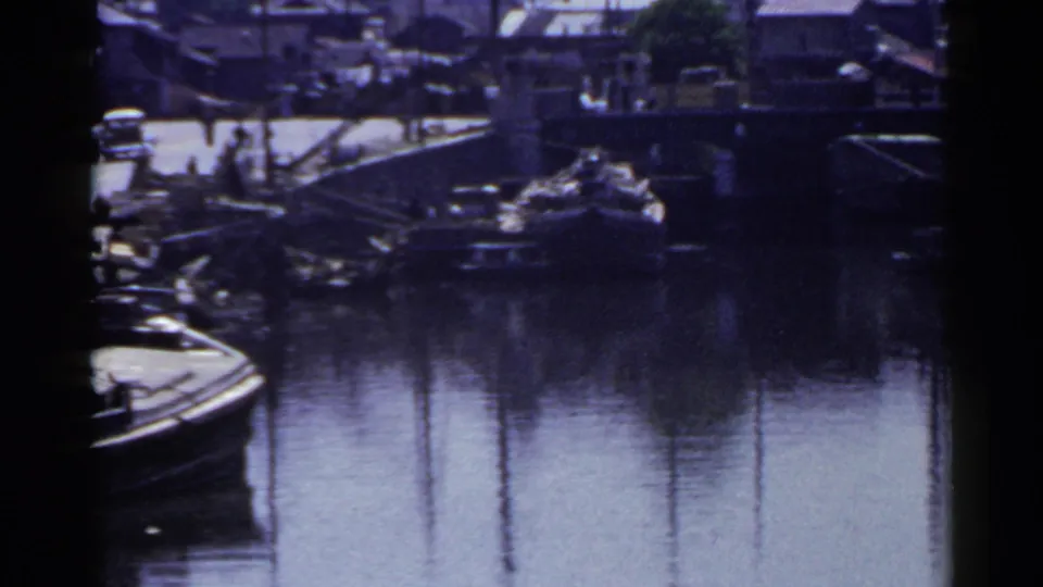 1953 Japan 8mm frame looking across a harbor canal toward docked boats, a bridge or ramp, small figures, and low buildings on the shore.