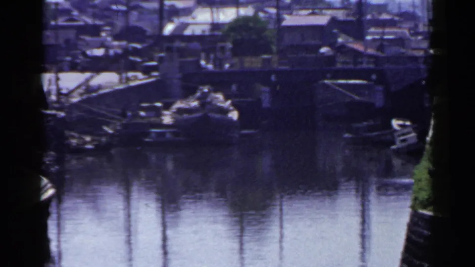1953 Japan harbor frame showing a broad area of reflective canal water with moored boats, vertical pole reflections, and waterfront buildings beyond.