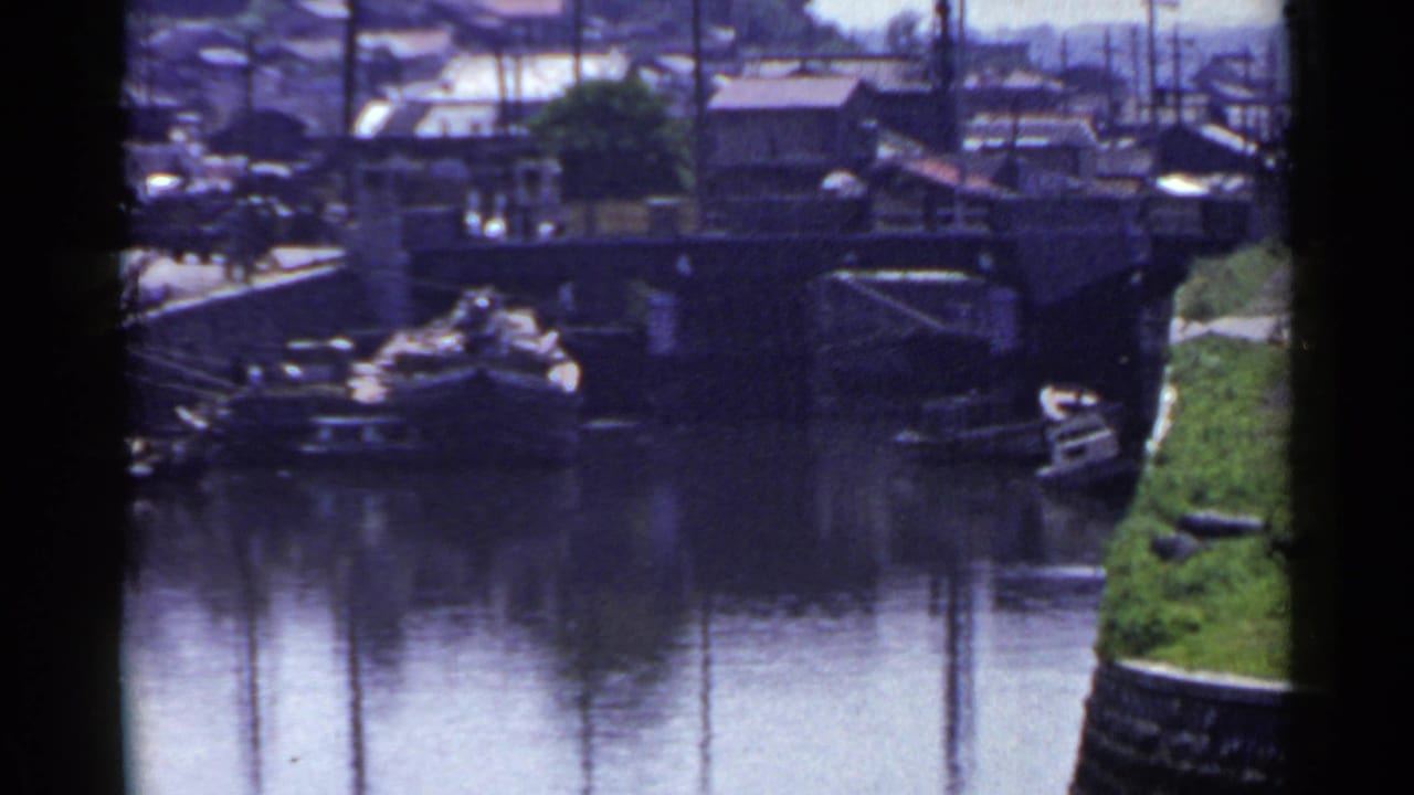 1953 Japan 8mm frame showing a canal view with bridge, moored boats, stone embankment, and shoreline buildings under clear daylight.