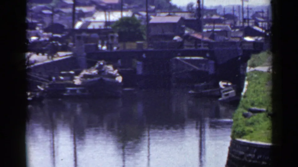 1953 Japan 8mm frame showing a canal view with bridge, moored boats, stone embankment, and shoreline buildings under clear daylight.