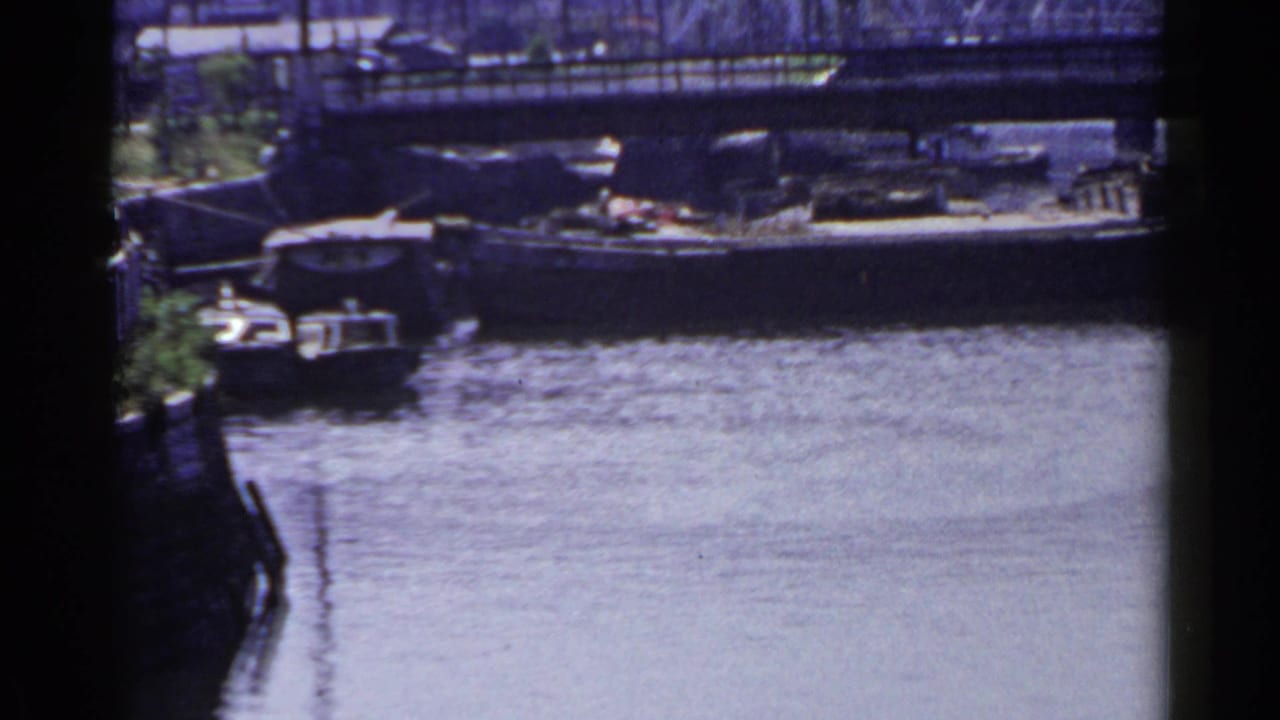 1953 Japan home-movie frame with a low bridge, calm water, a moored boat, and distant waterfront structures.