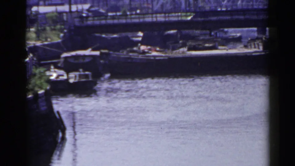 1953 Japan home-movie frame with a low bridge, calm water, a moored boat, and distant waterfront structures.