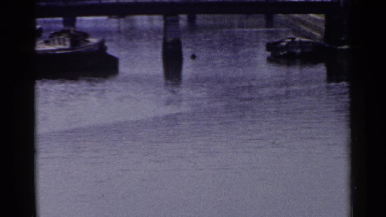 1953 Japan harbor view with calm water, dark structural beams, small boats, and stone-lined waterfront edges.