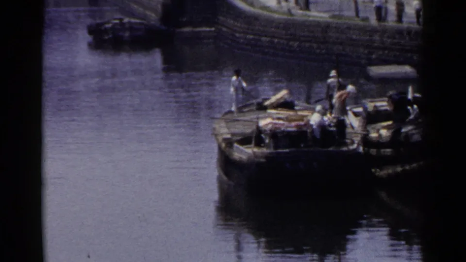 In 1953 Japan, people stand on or near a moored wooden boat along a quiet stone-lined harbor edge.