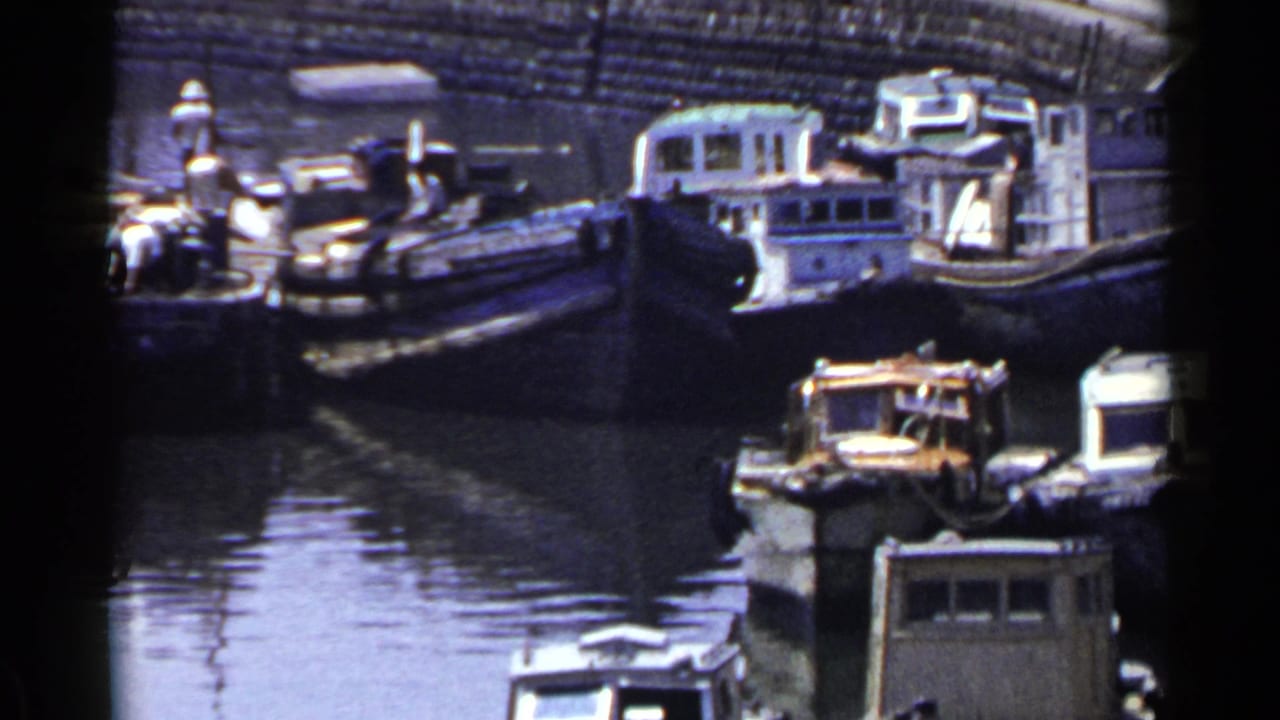 Small boats moored along a canal with vehicles and pedestrians near a stone embankment.