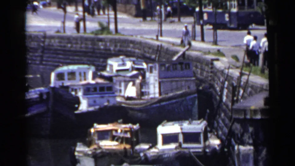 1953 Japan frame showing a marina below a street, with pedestrians, vehicles, stonework, and small boats sharing the waterfront view.