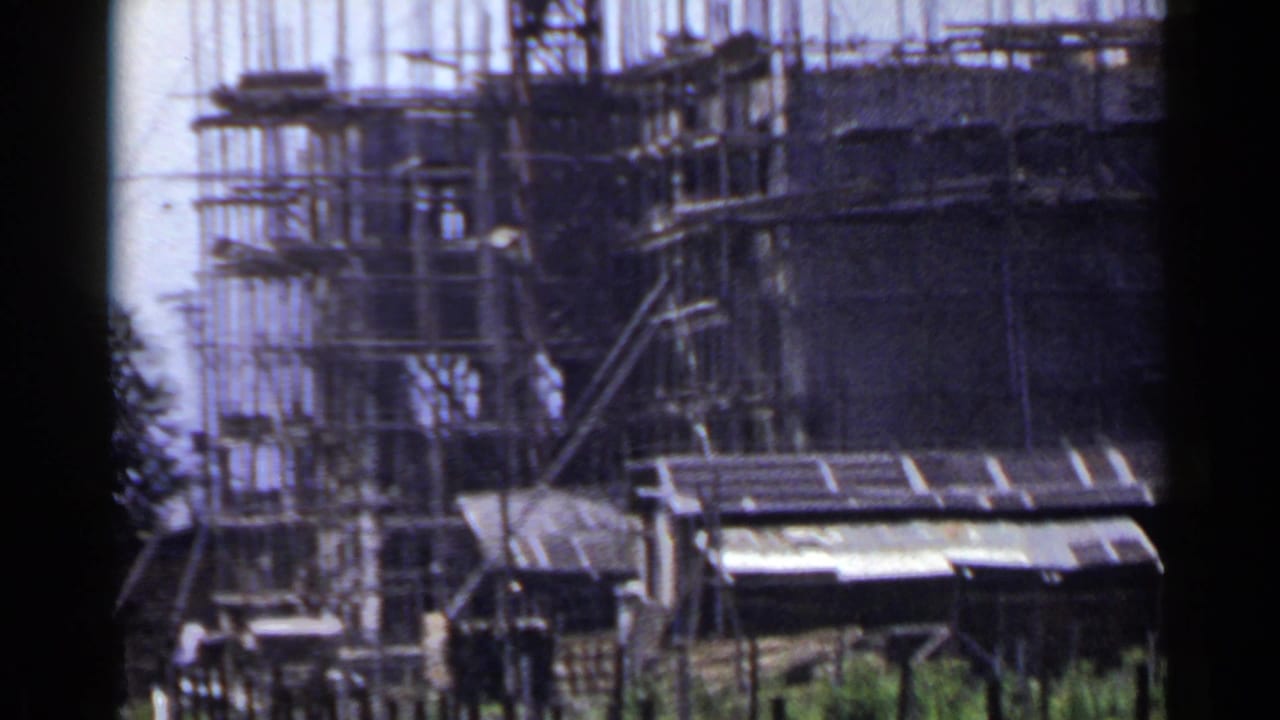 1953 Japan 8mm frame showing a mid-rise construction site with scaffolding, vertical rebar, sheds, and small workers near the base.