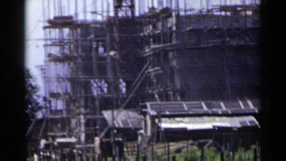 1953 Japan 8mm frame showing a mid-rise construction site with scaffolding, vertical rebar, sheds, and small workers near the base.