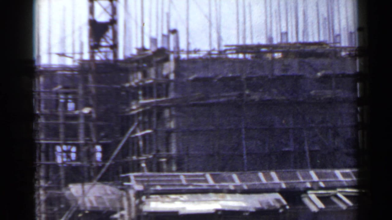 1953 Japan home-movie frame of a partially built mid-rise with scaffolds, rebar, and a temporary shed in the foreground.
