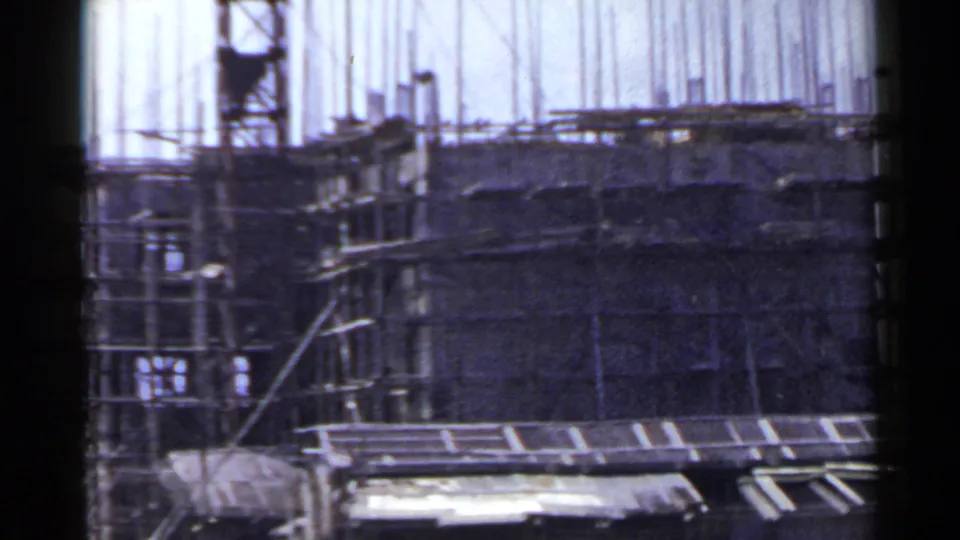 1953 Japan home-movie frame of a partially built mid-rise with scaffolds, rebar, and a temporary shed in the foreground.