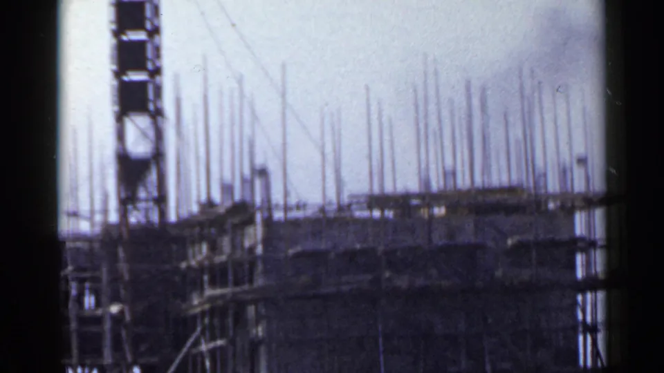1953 Japan 8mm frame showing a vertical hoist or tower next to a scaffolded mid-rise with rebar rising above the roofline.