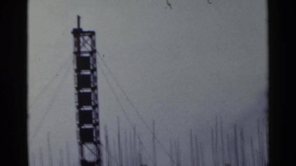 1953 Japan home-movie frame with a tall hoist-like tower and thin rebar lines silhouetted above the construction site.