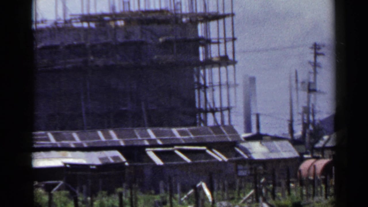 1953 Japan 8mm frame showing a scaffolded concrete building behind low corrugated-roof sheds and utility poles.