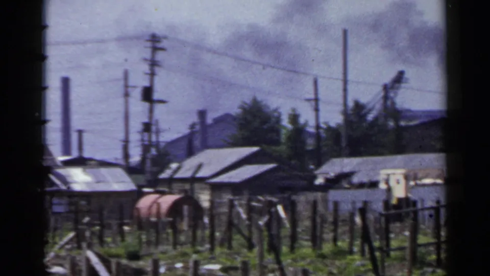 1953 Japan 8mm frame showing low industrial buildings, rough fencing, utility poles, and distant smoke.