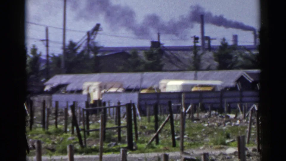 1953 Japan 8mm frame showing fenced industrial ground, low sheds, distant chimneys, and smoke drifting across the sky.
