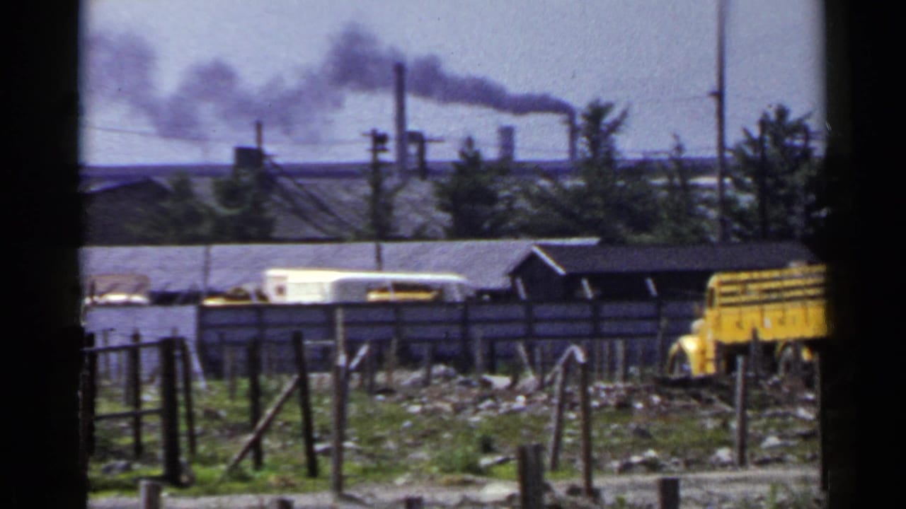 1953 Japan 8mm frame showing factory smoke beyond fences, utility poles, debris, and a yellow truck in an industrial yard.