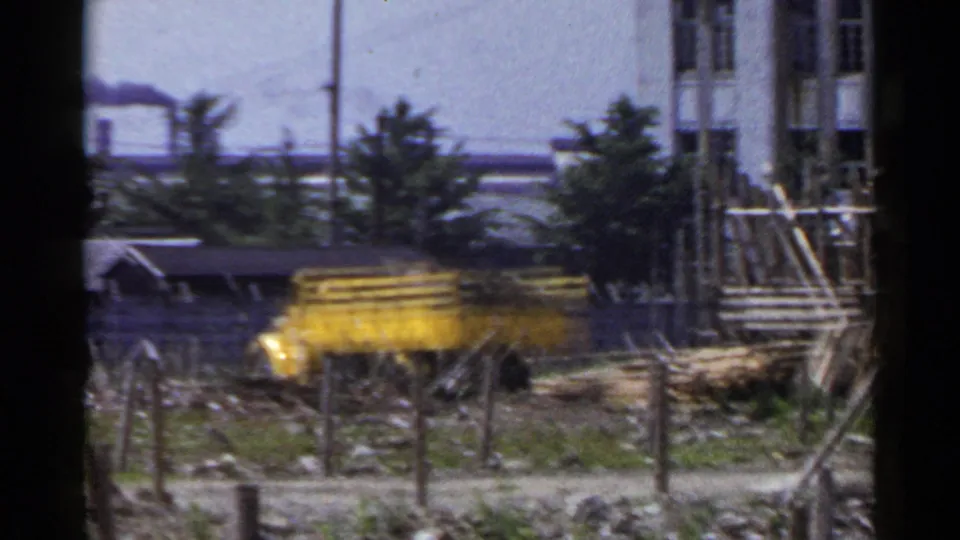 Yellow dump truck near a rough construction area in Japan, 1953, with smoke drifting from an industrial pipe behind it.