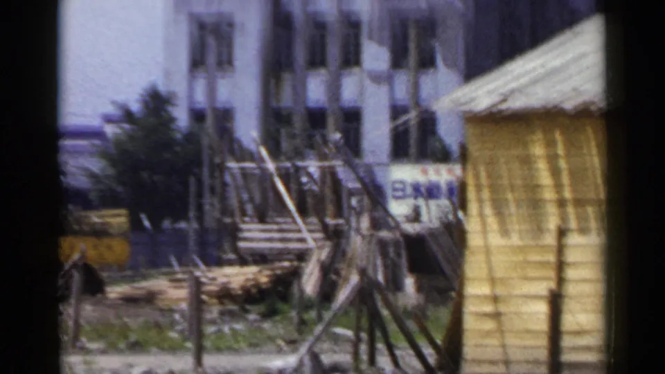 1953 Japan home-movie frame showing wooden scaffolding, fence posts, a pale building, and partial Japanese signage near a worksite.