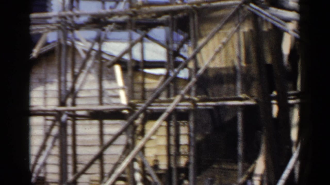 1953 Japan home-movie frame showing dense bamboo scaffolding and diagonal braces against a wooden building exterior.