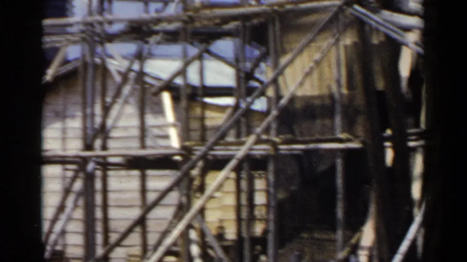 1953 Japan home-movie frame showing dense bamboo scaffolding and diagonal braces against a wooden building exterior.