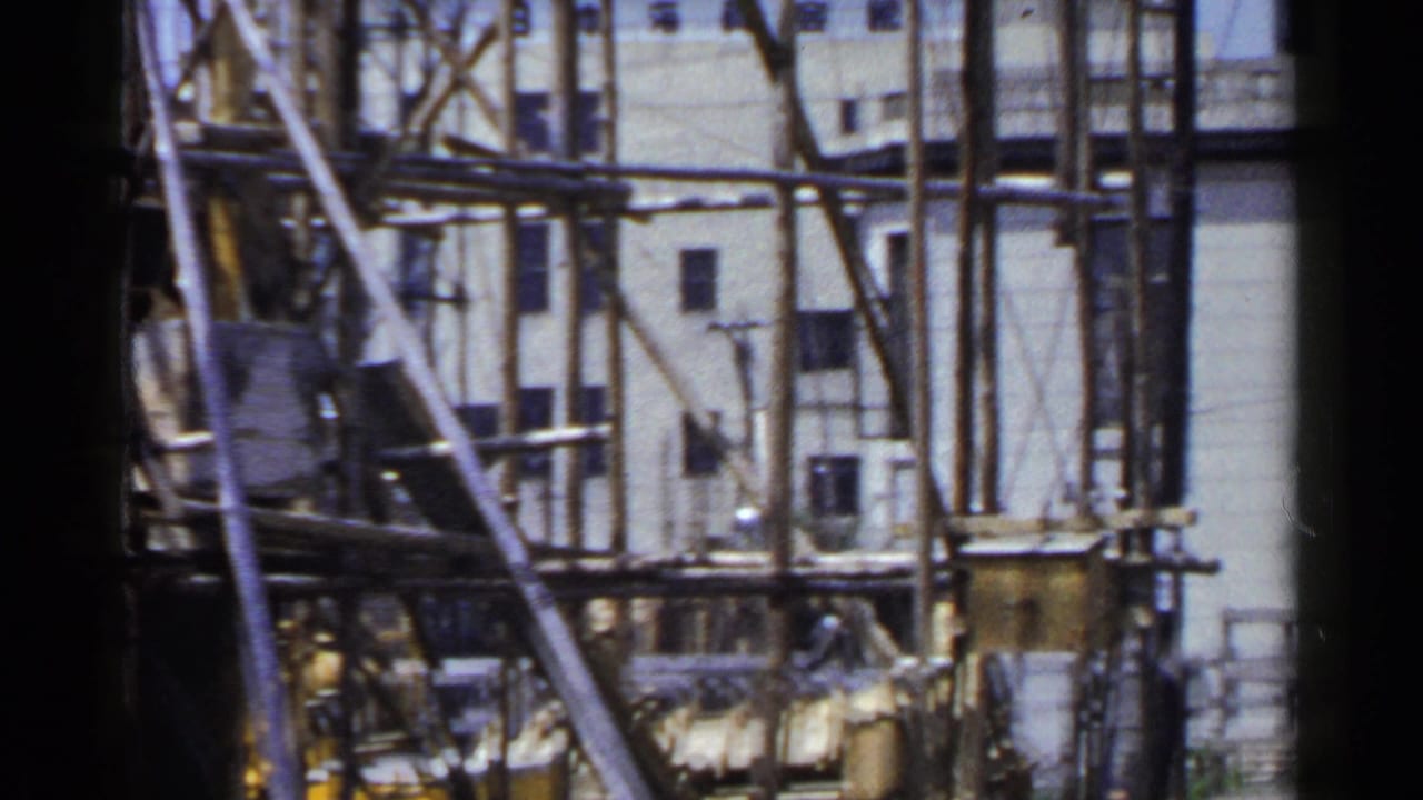 1953 Japan home-movie frame with dense wooden scaffold poles crossing a pale exterior wall with small windows.